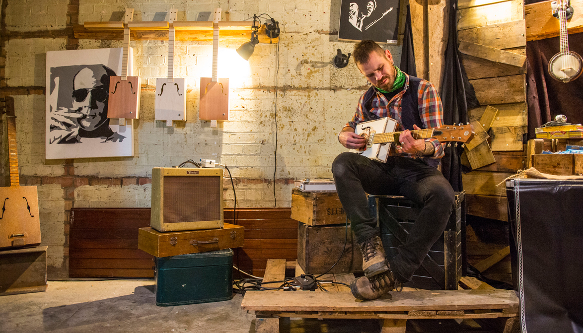 May playing a tissue box guitar he has made, sat on wooden crates