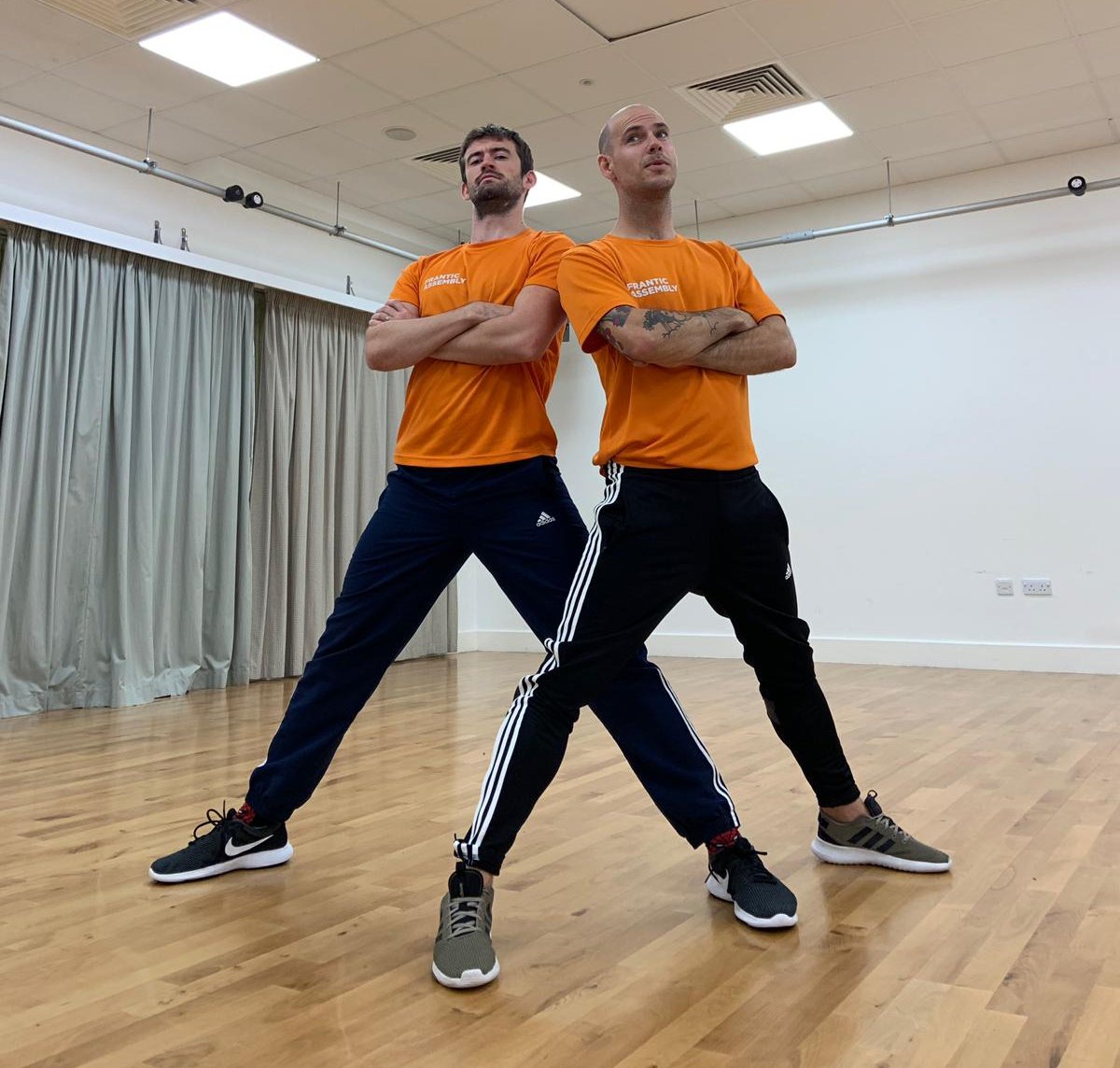 two men standing with their arms crossed in a rehearsal space.