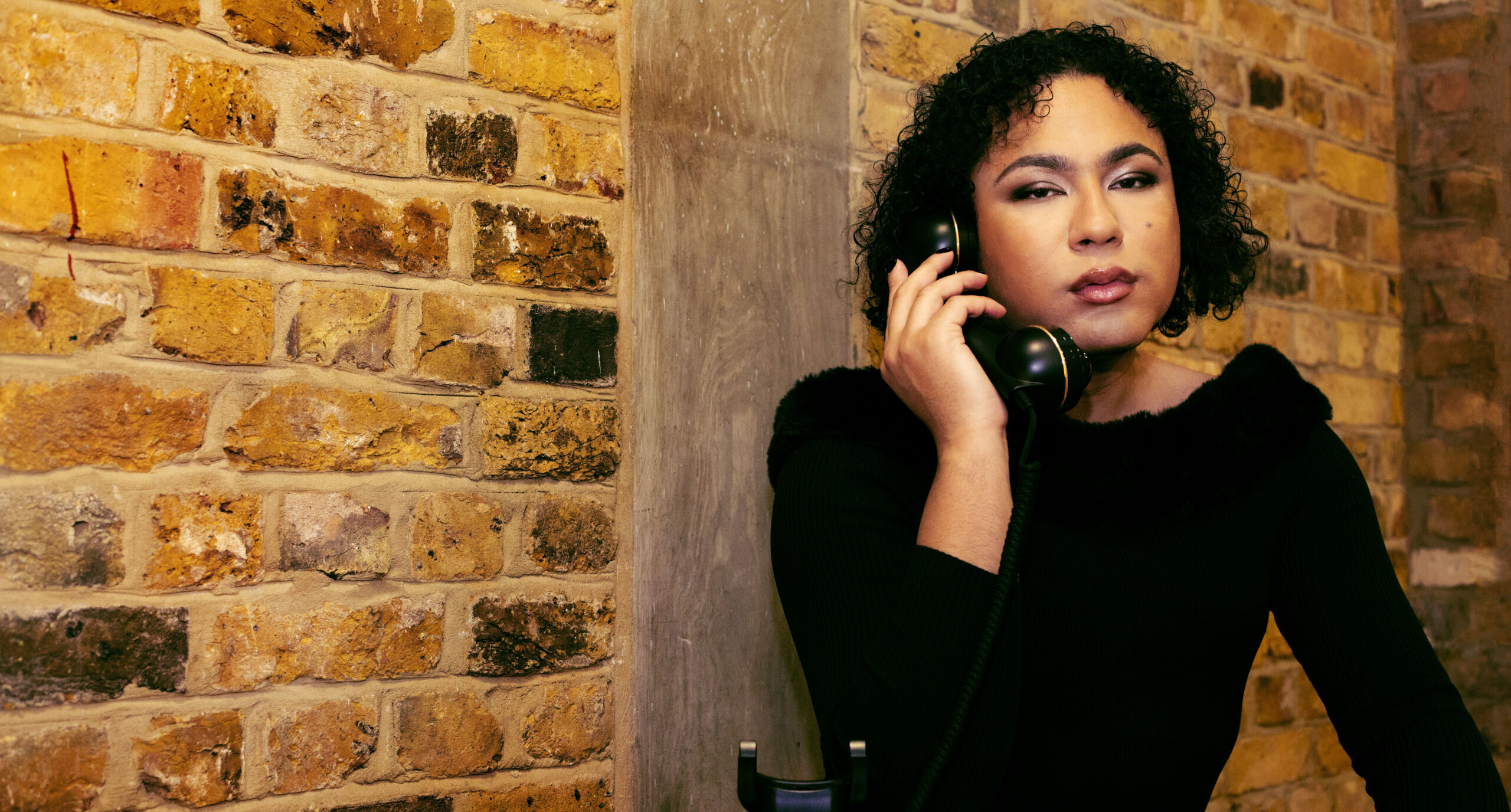 A person dressed in black sits looking at the camera, holding a black landline phone to their ear