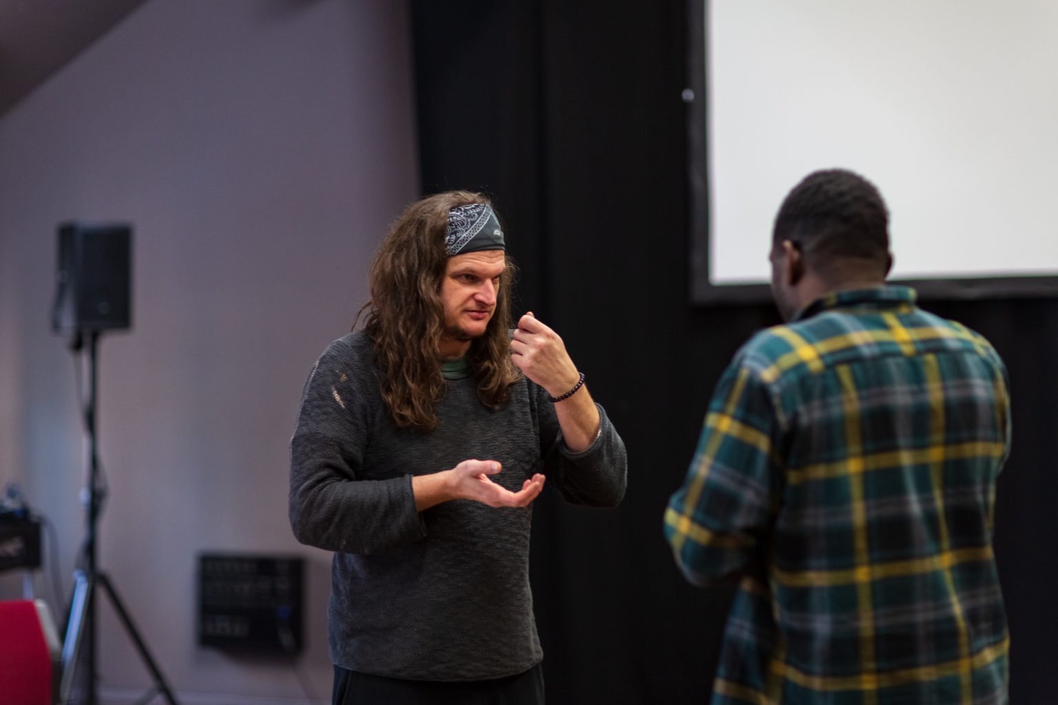 A white man stands in the middle of a rehearsal room. He has long brown hair, and wears a dark green head band with white patterns. He has a dark green jumper on and holds his hands up in front of his body. He is giving feedback in BSL to a black male performer, who has his back to the camera and wears a green and yellow plaid shirt.