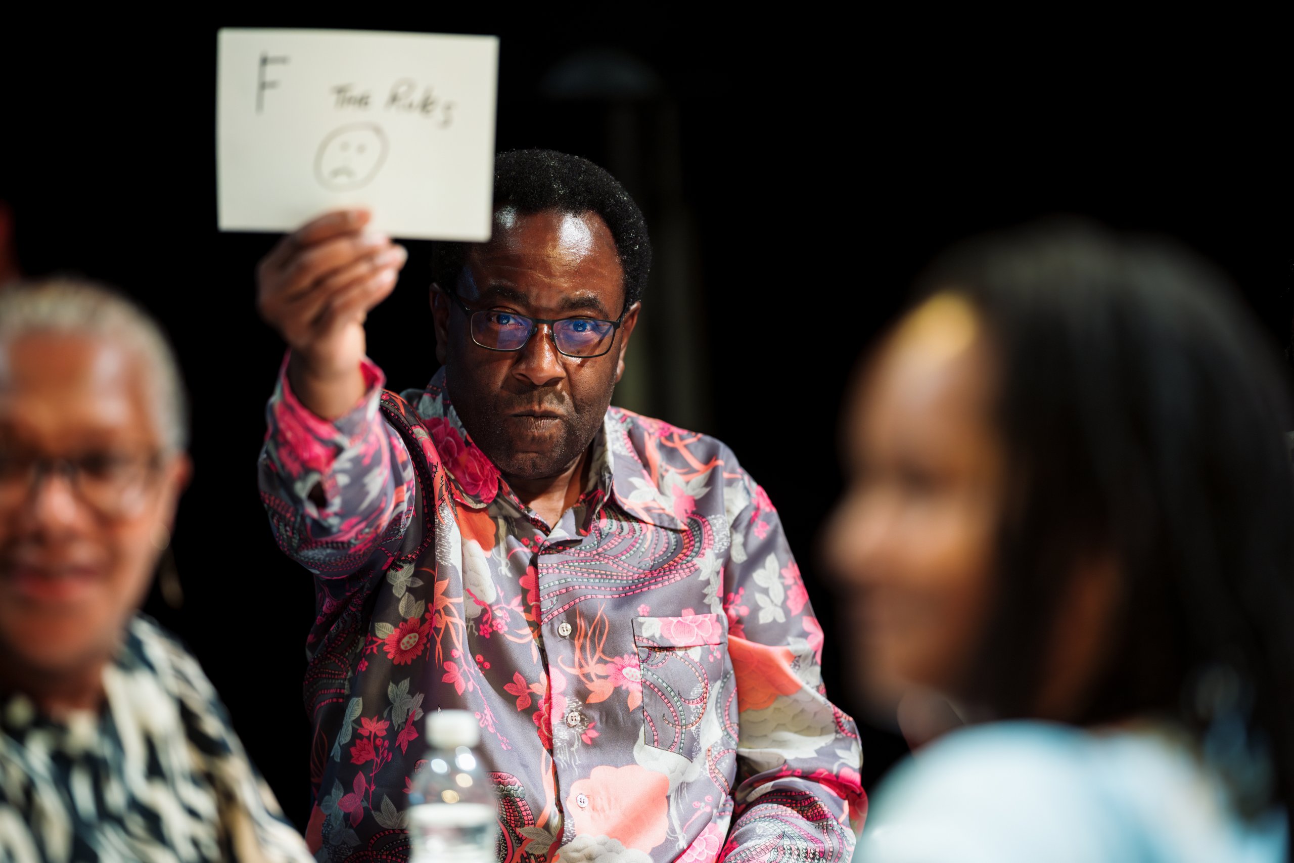 A black male, aged 40s-50s, playing 1884 at Fairfield Halls, Croydon. He is wearing a colourful, patterned shirt and looking directly at the camera with a determined facial expression. He is holding up a protest sign with ‘F the rules’ and a sad face written on it.
