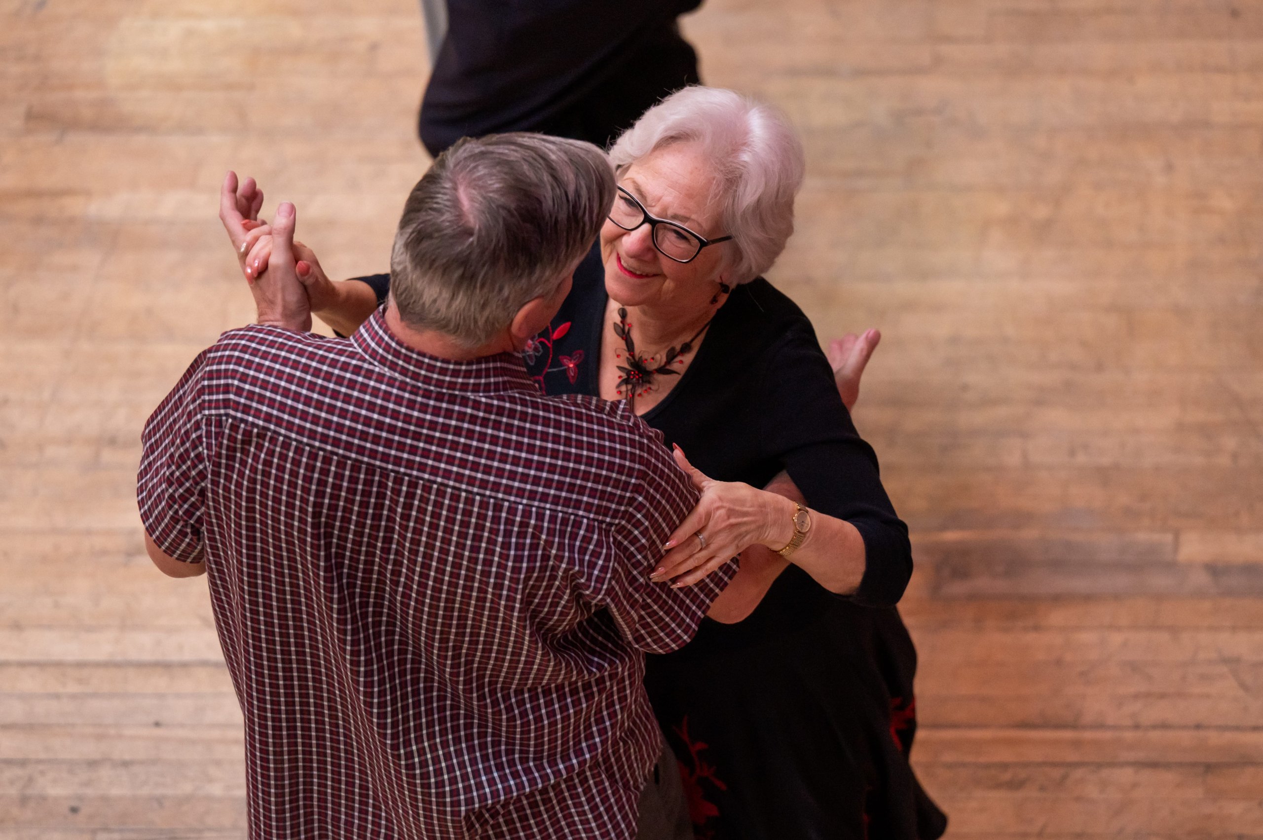 Two Tea Dancers stand in a ballroom hold position, smiling at each other. It is a sweet and intimate moment with a hint of festive purple lighting shining down on them. It's cosy