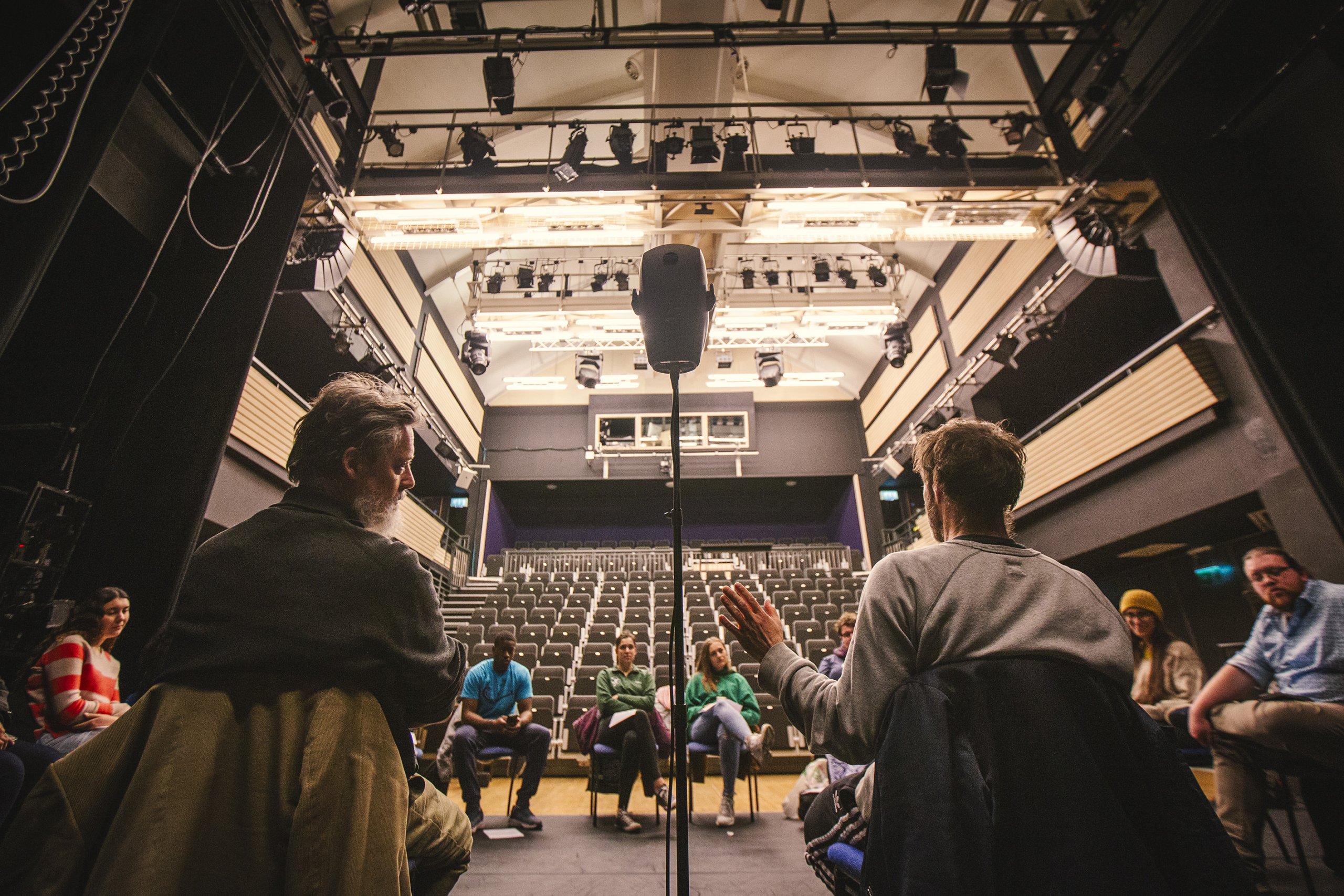 The camera looks out from the back of a theatre stage - a group of participants sit in a circle on stage with the audience seats dramatically in view in the background. Two DARKFIELD team members sit in view facing the participants, speaking to them. In the center of the image and between the two team members, there stands a binaural head microphone on a microphone stand, as tall as a person.